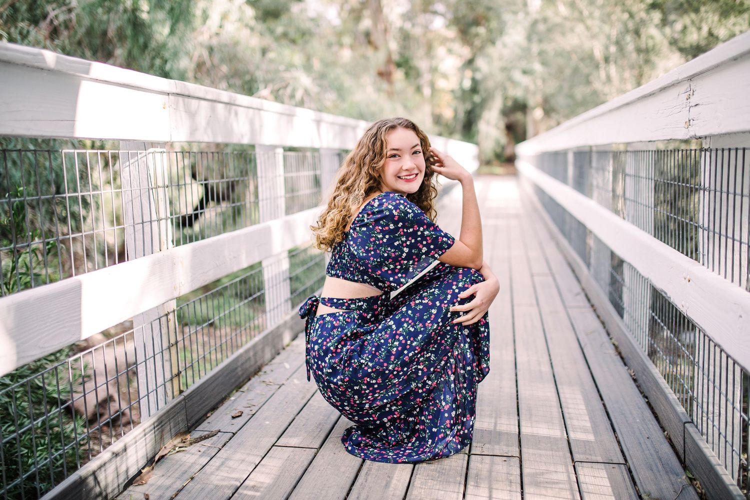 Smiling young woman in floral dress crouching on a wooden bridge surrounded by lush greenery.