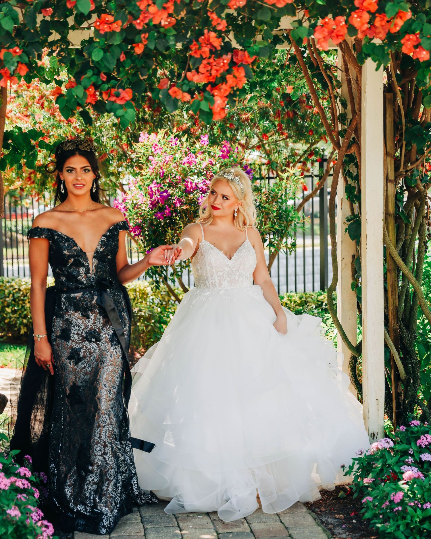 Two elegant gowns, one black lace and one white tulle, photographed in a garden setting with vibrant coral flowers.