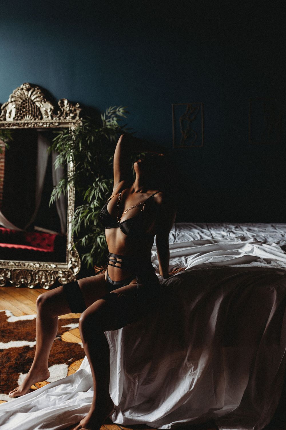 Figure in black lingerie posed dramatically on bed with ornate mirror in background.