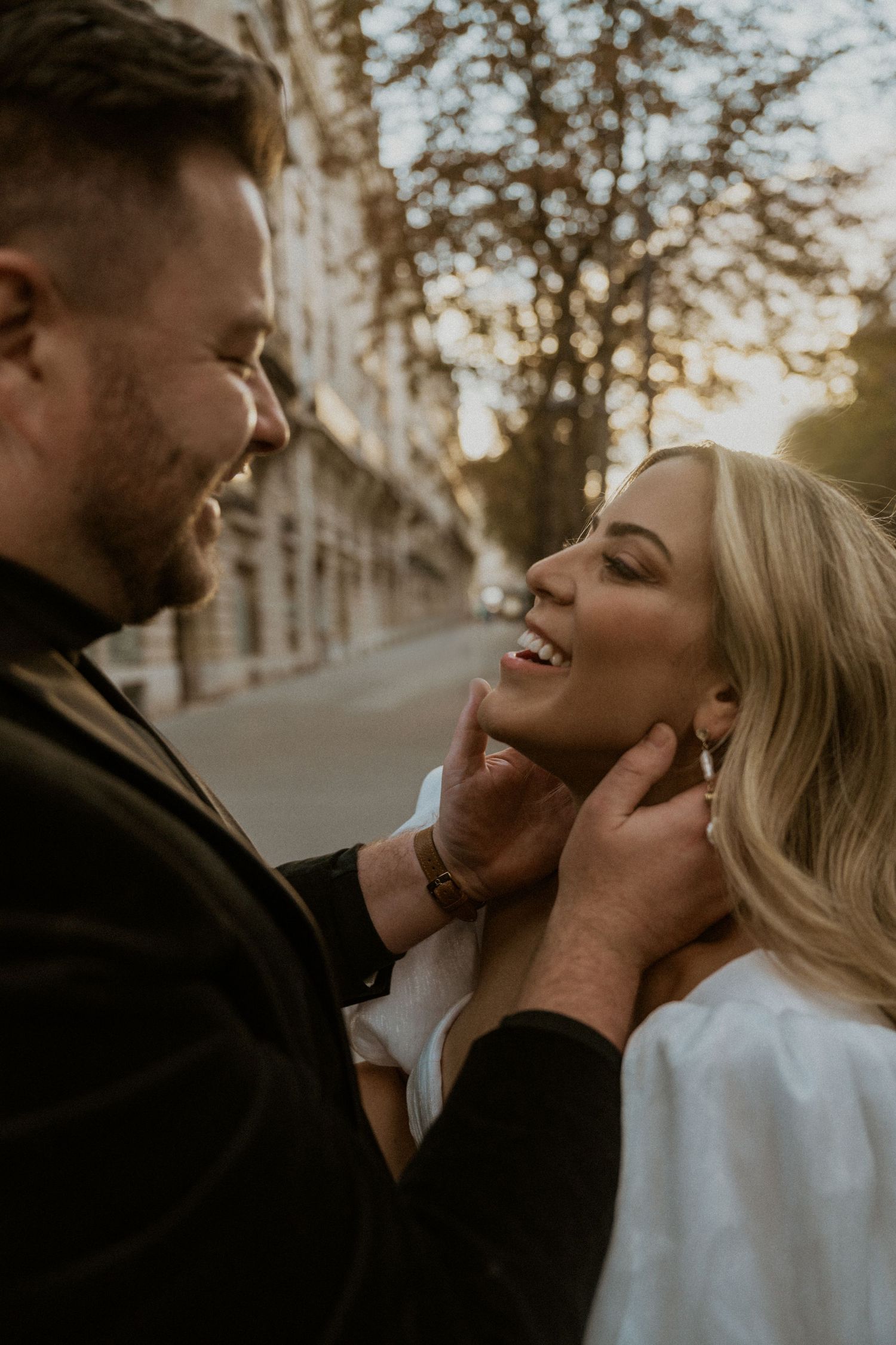 Romantic couple sharing an intimate moment outdoors during golden hour with tree-lined street in background.