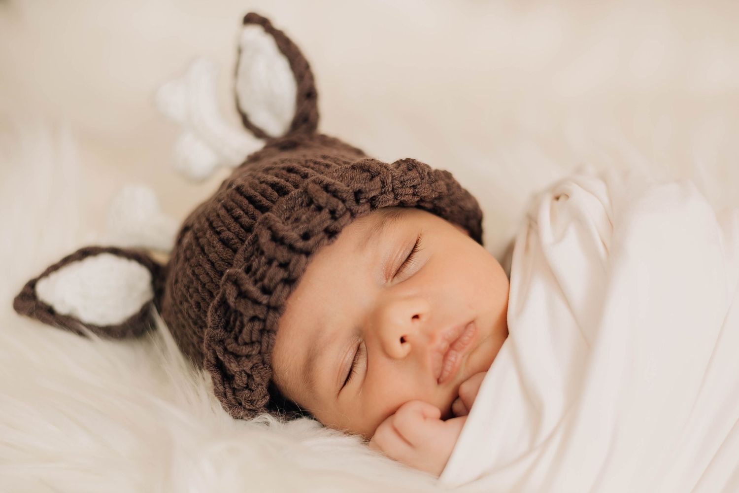 Sleeping newborn baby wearing a brown knit deer hat with ears, swaddled in white.