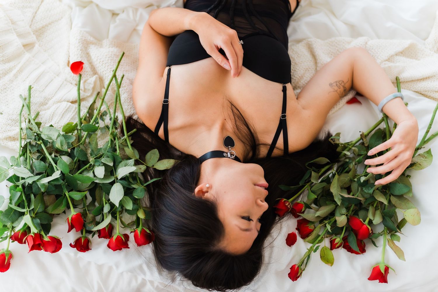 Person lying on white bedding surrounded by red roses and greenery in artistic portrait.