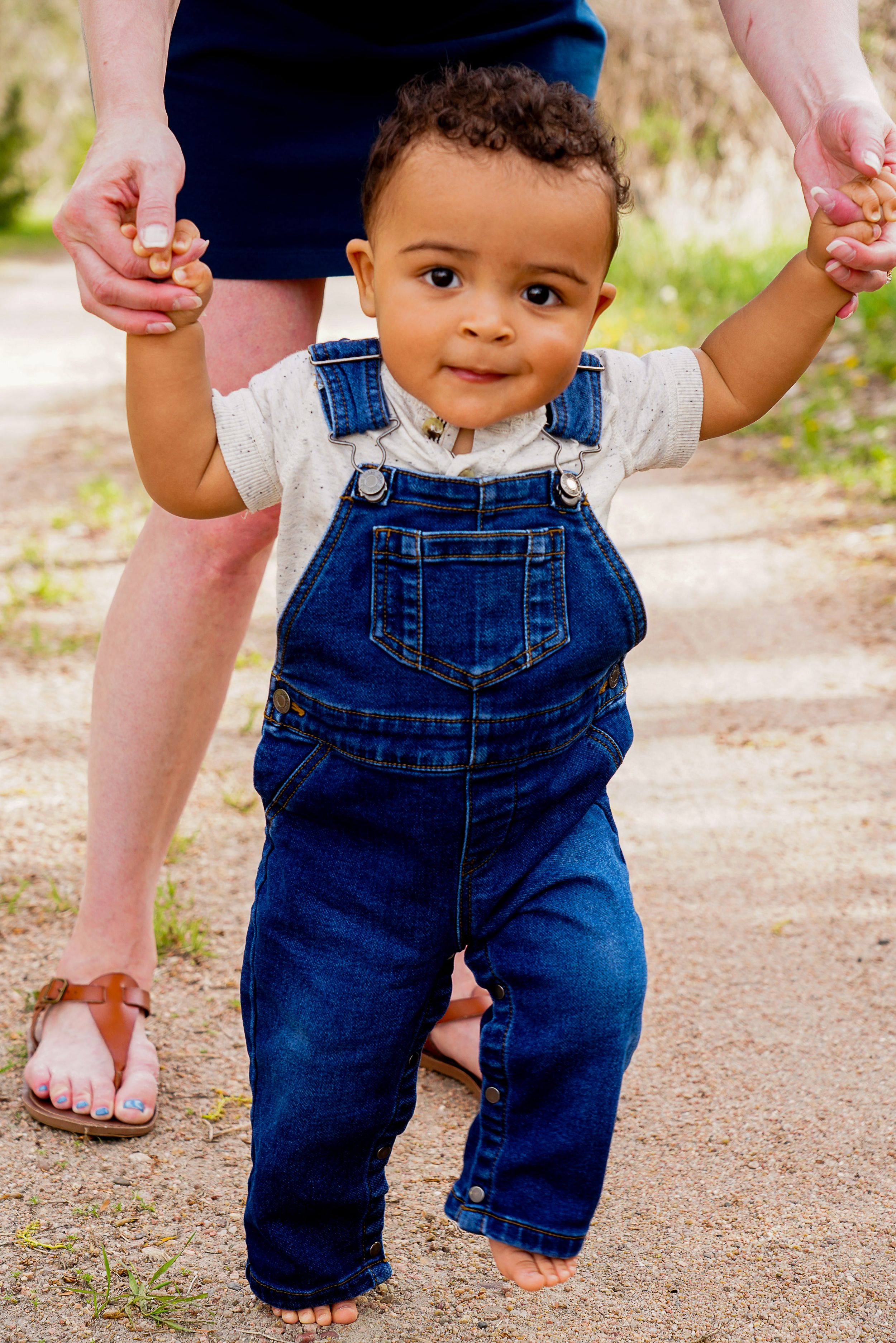 Toddler in denim overalls holding hands while learning to walk on a dirt path.