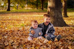 Two children sitting and laughing in pile of autumn leaves under tree.