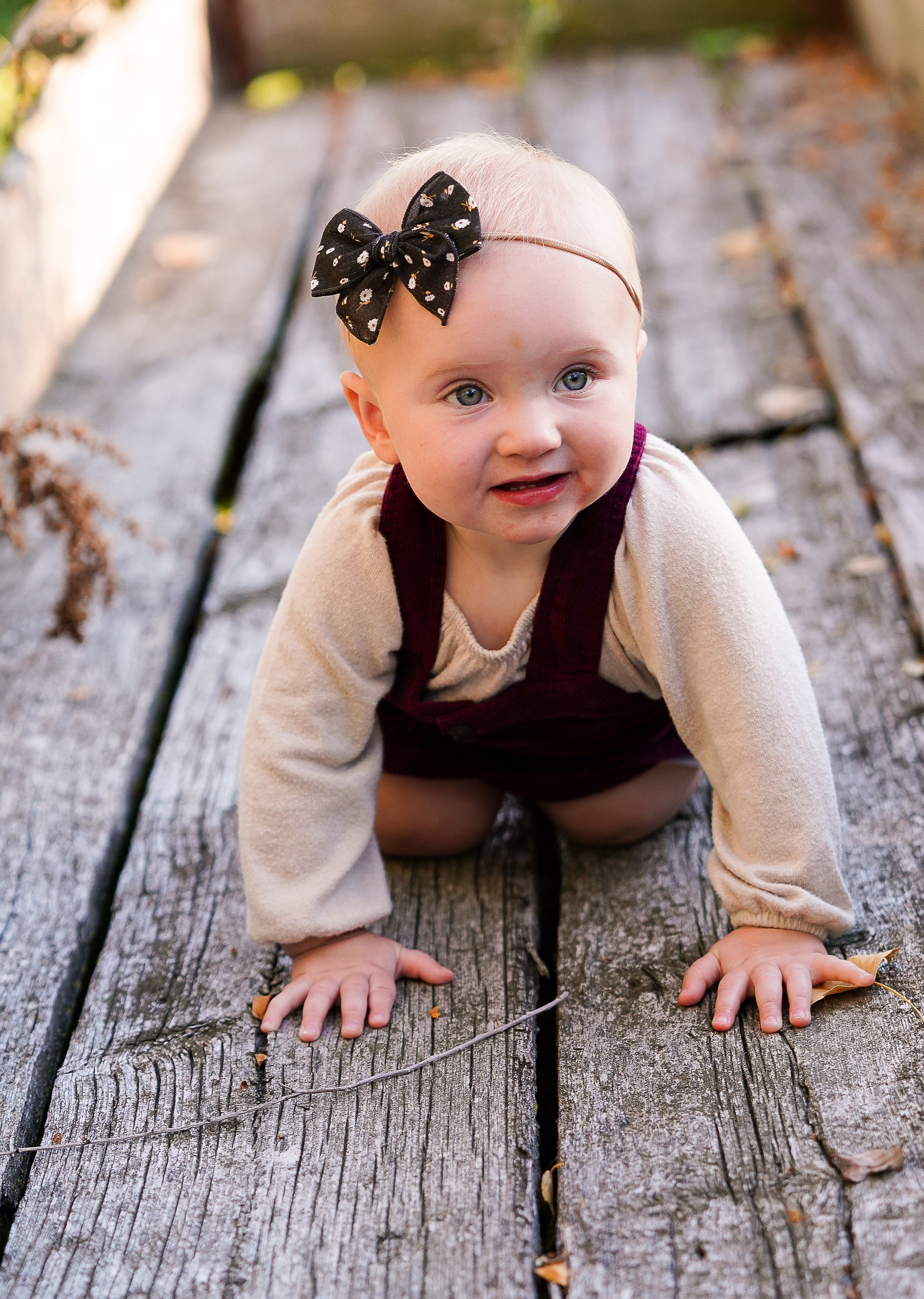 A baby in a brown outfit with polka dot bow headband crawls on wooden decking.