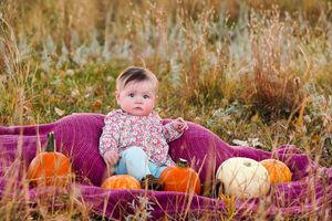 Baby sits on purple blanket surrounded by orange and white pumpkins in autumn field.