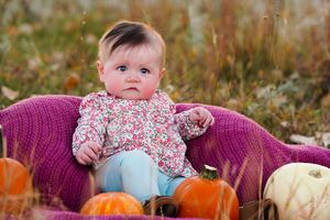 Baby in floral outfit sits on purple blanket with orange and white pumpkins.