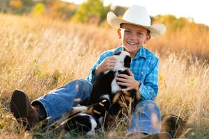 Young person in cowboy hat and denim holds black and white puppy in golden field.