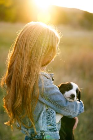 Person with long blonde hair holds puppy while standing in golden sunset field.