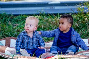 Two toddlers in blue shirts sit together on patterned blanket outdoors.