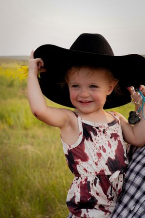 A child in a floral dress playfully poses outdoors while wearing an oversized black cowboy hat.