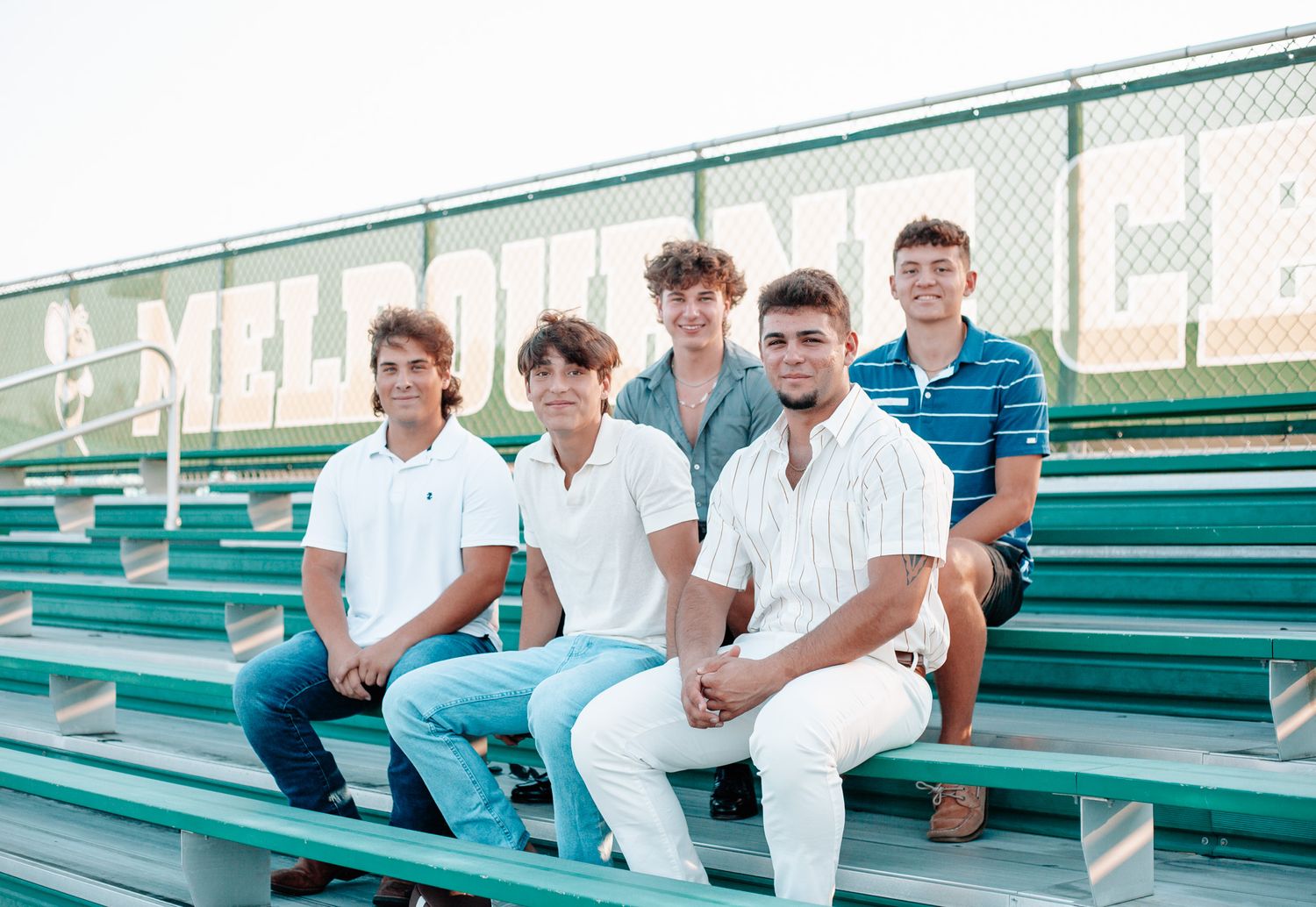 Group of students in white shirts sitting on green bleachers at a sports venue.