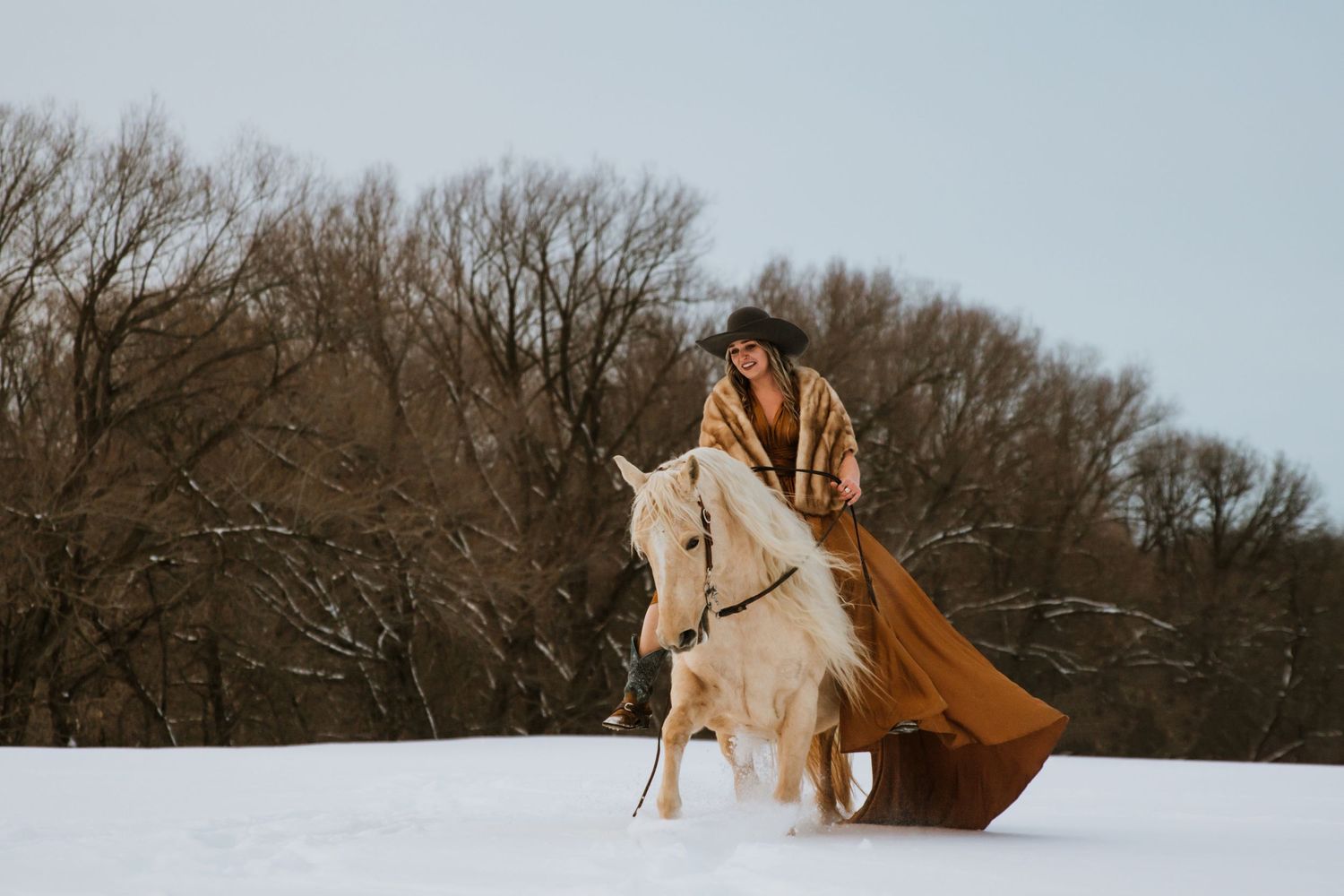 A horse and rider in flowing rust-colored clothing gallop gracefully through a snowy landscape.