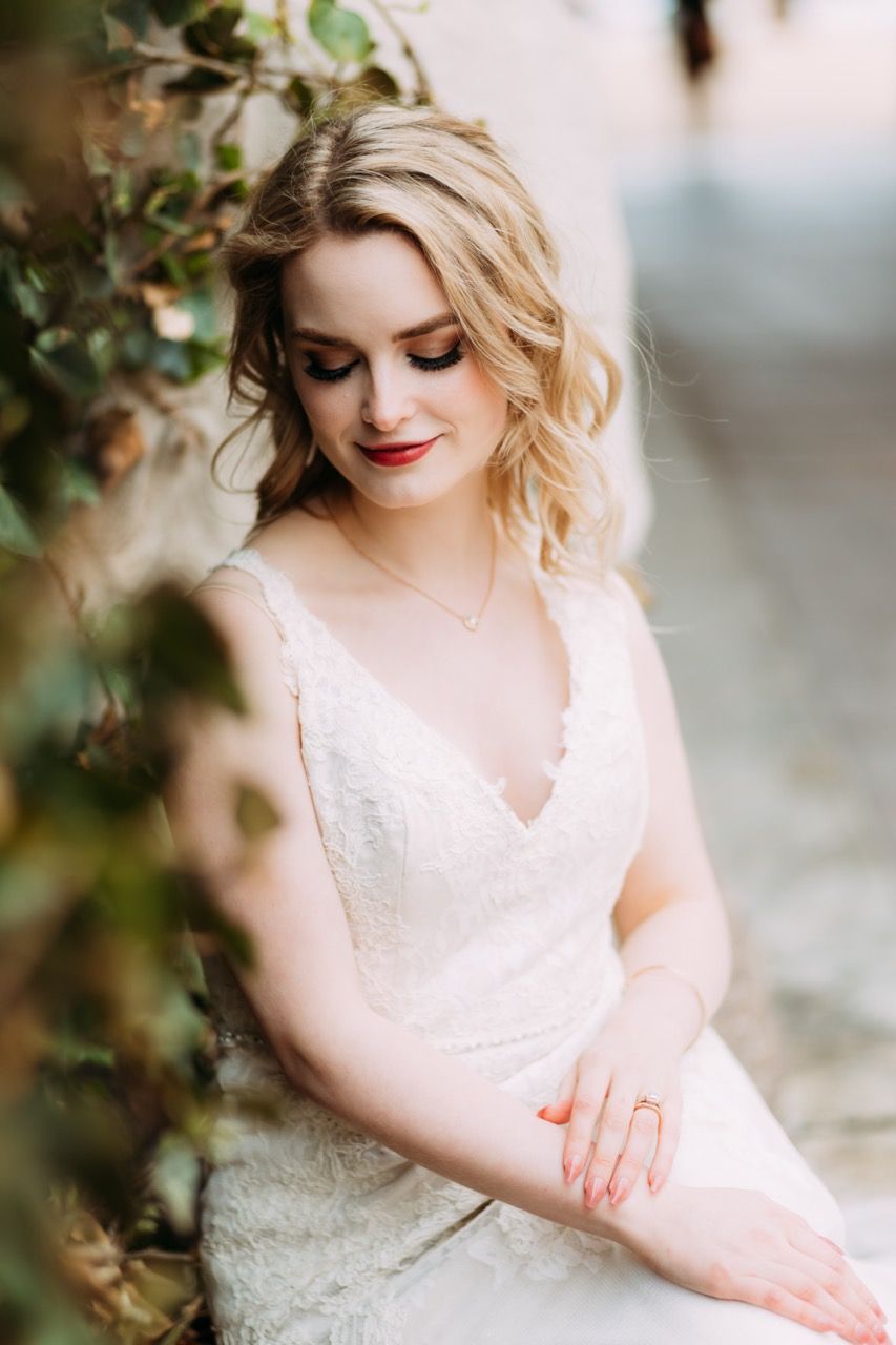 A blonde woman in a white lace dress sits with her hands gently resting on her lap. She is looking down and smiling soft