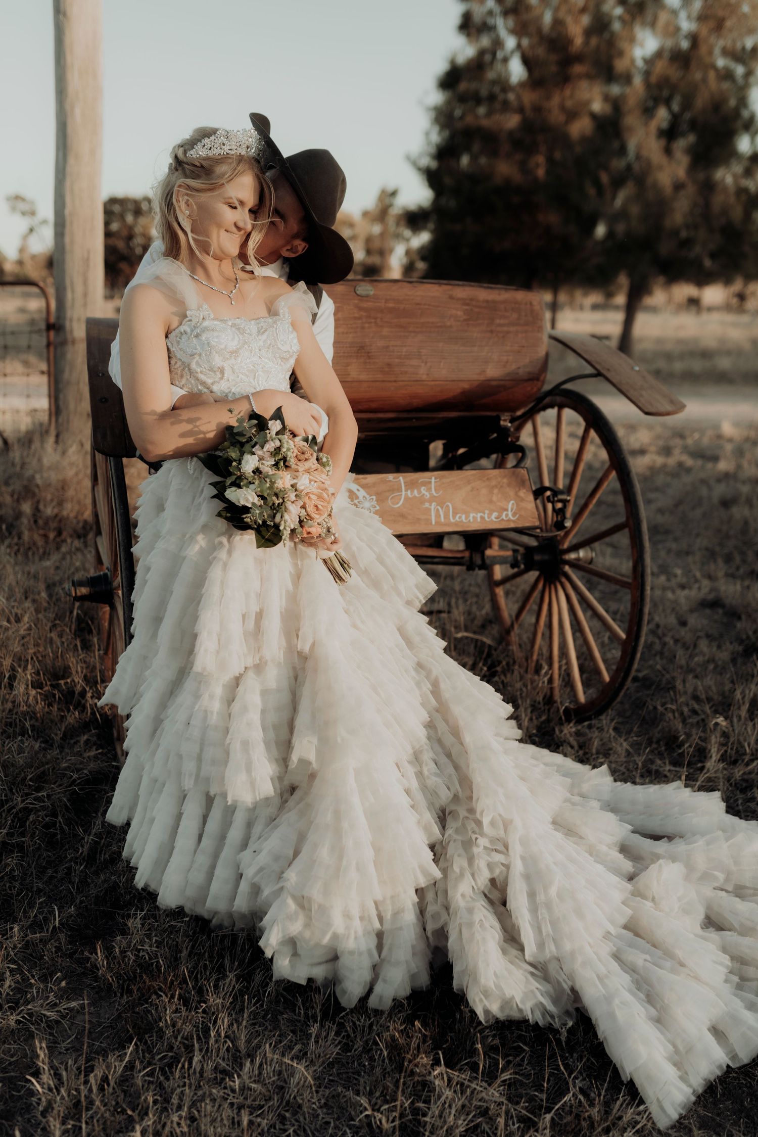 A bride in a flowing white dress poses near a vintage wooden wagon at sunset.