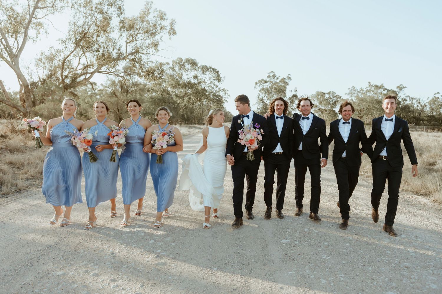 Wedding party in blue and black attire walking together on a rural path.
