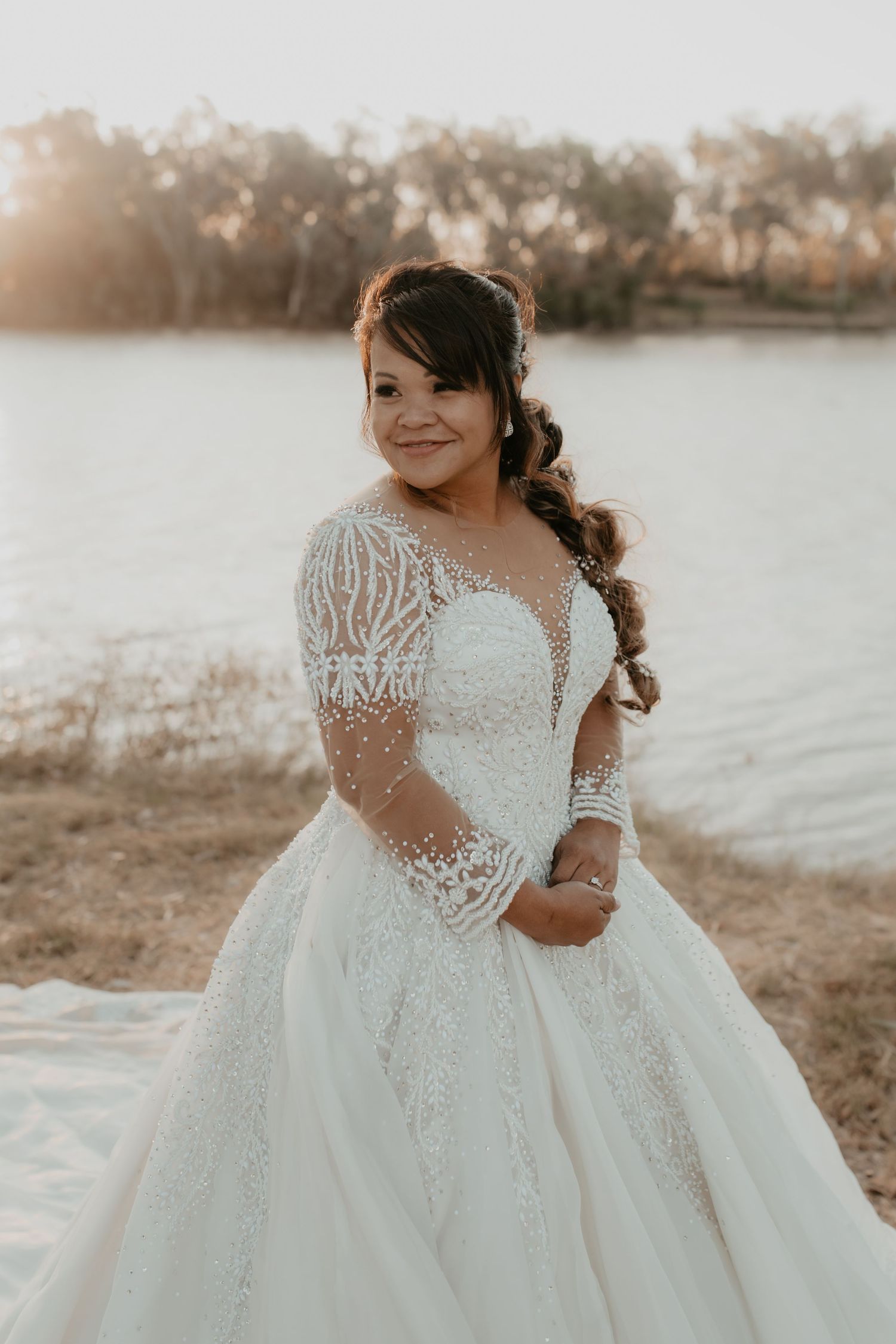 A bride poses at sunset by a lake wearing an elegant lace ball gown wedding dress.