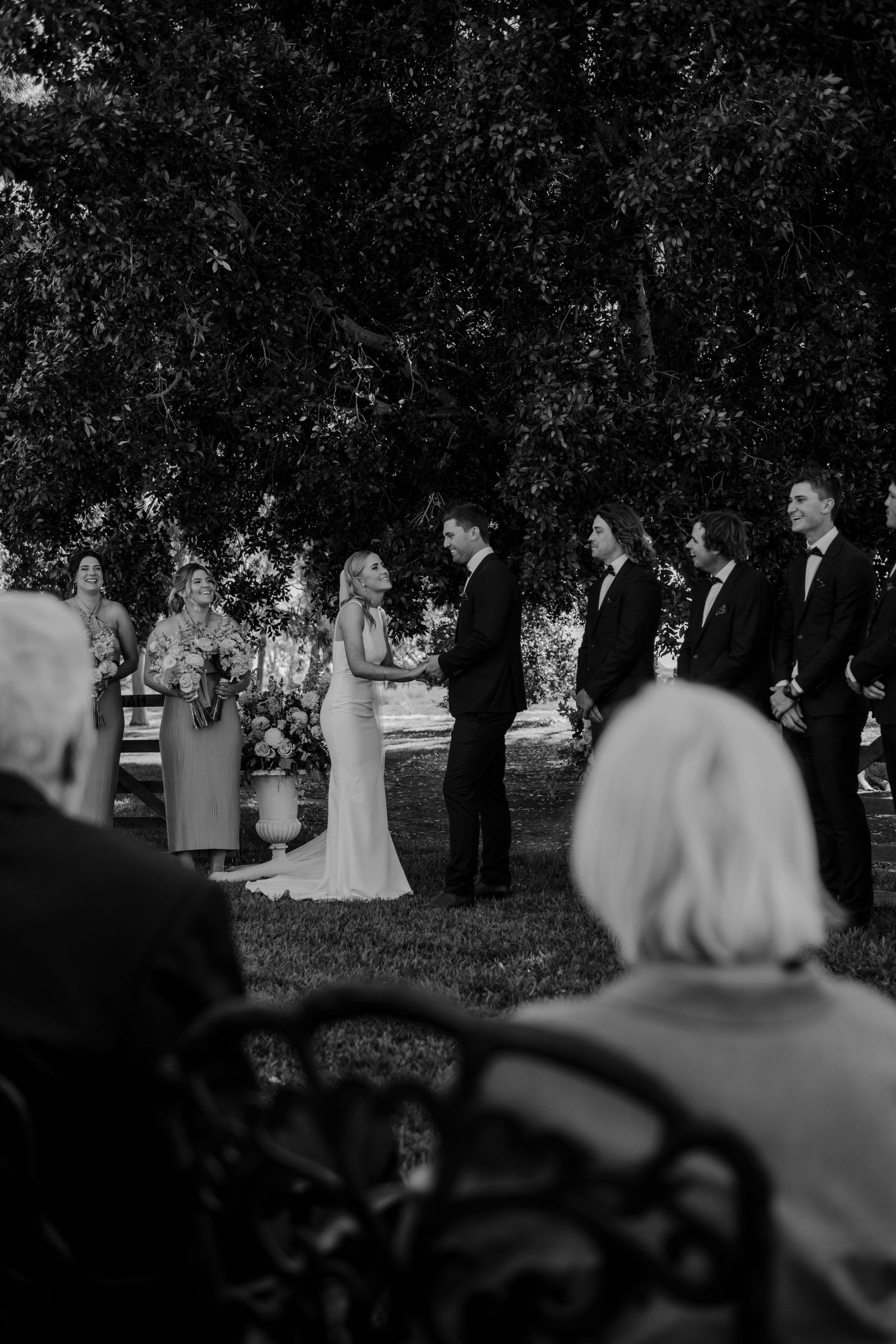 A black and white outdoor wedding ceremony under trees with guests watching.