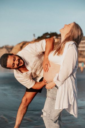 Playful maternity photo of couple laughing together on beach at sunset.