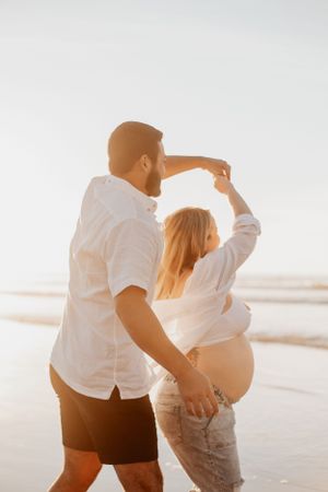 Expecting couple dancing together on a sunlit beach during their maternity photoshoot.