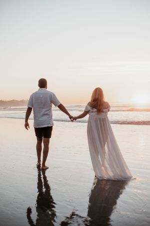Couple holding hands walking through ocean waves at sunset on the beach.