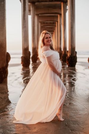 Maternity photo of woman in white dress under pier during golden hour.
