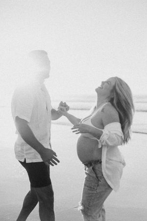 Black and white photo of an expecting couple sharing a playful moment on the beach.