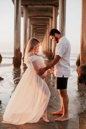 Couple sharing intimate moment under pier during sunset maternity photoshoot.