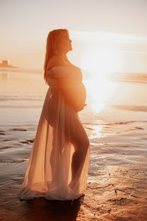 Silhouette of pregnant woman in flowing dress standing on beach at sunset.