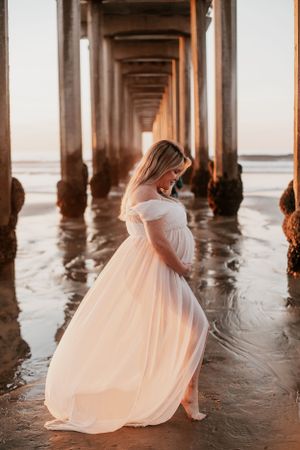 Side view of pregnant woman in flowing white dress under oceanside pier.