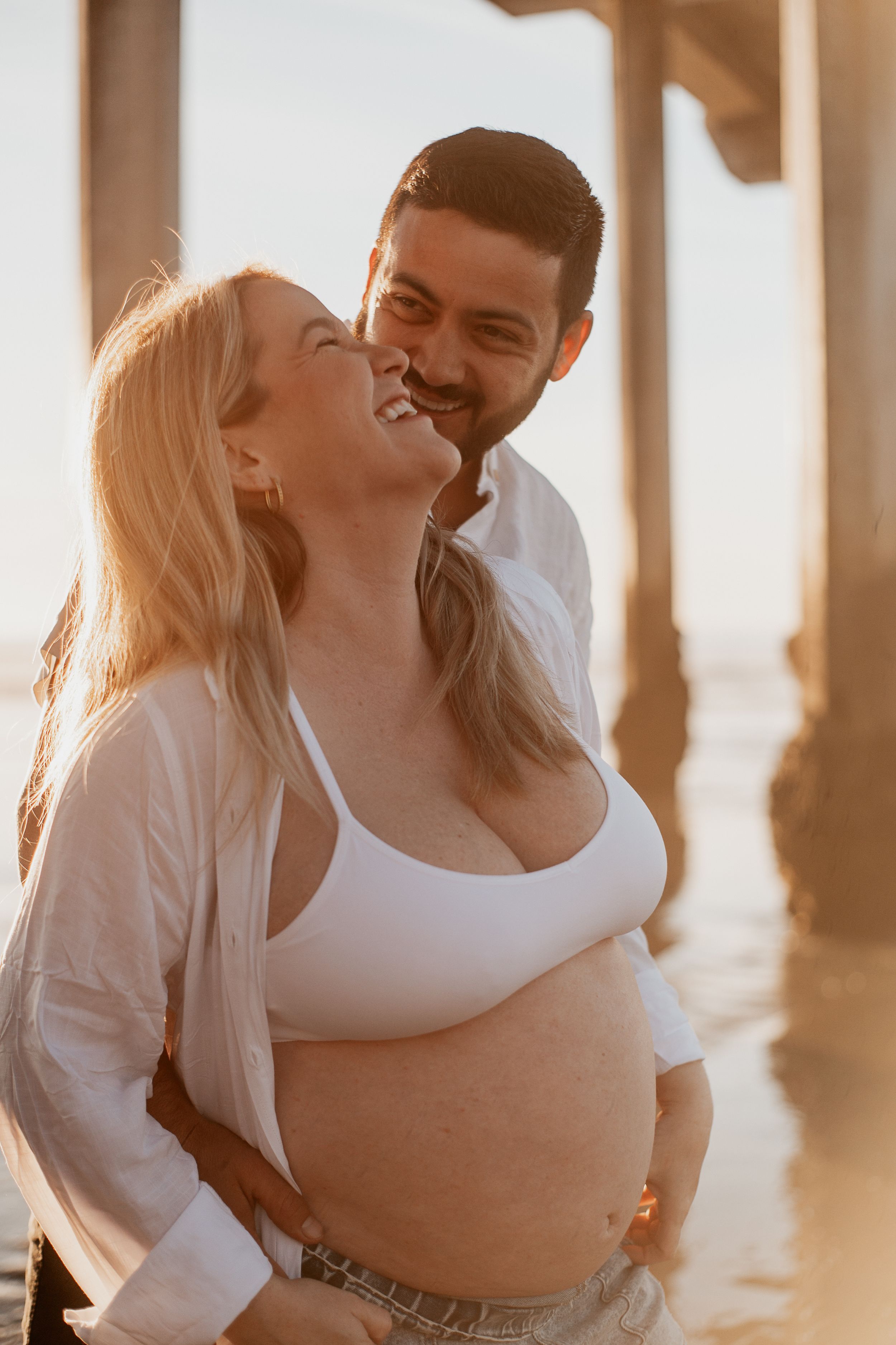 Joyful maternity moment captured between couple on beach pier at sunset.