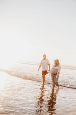 Couple walking through shallow ocean water during sunset maternity photoshoot.