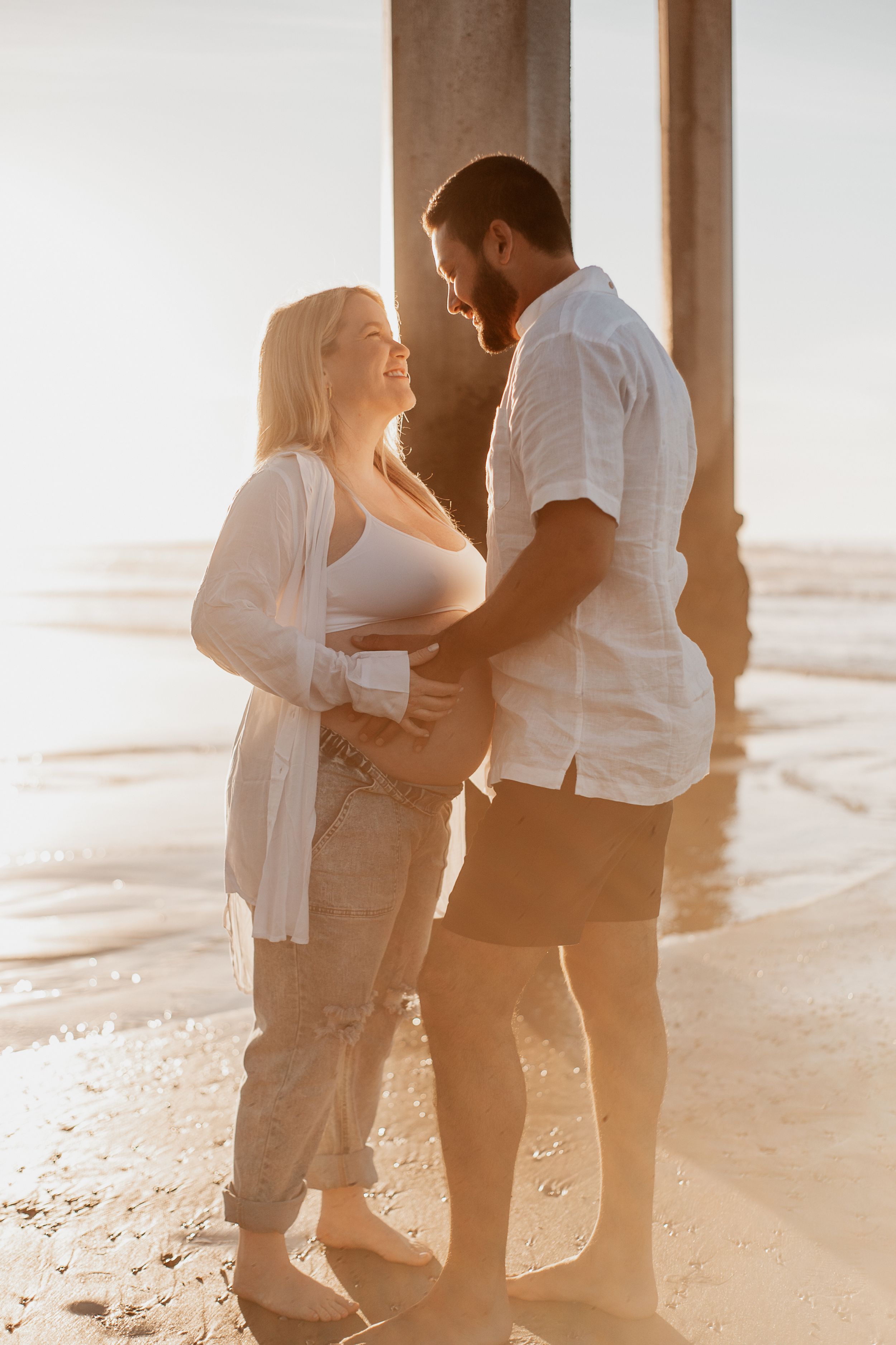 Romantic beach maternity photo of couple in white embracing at golden hour.
