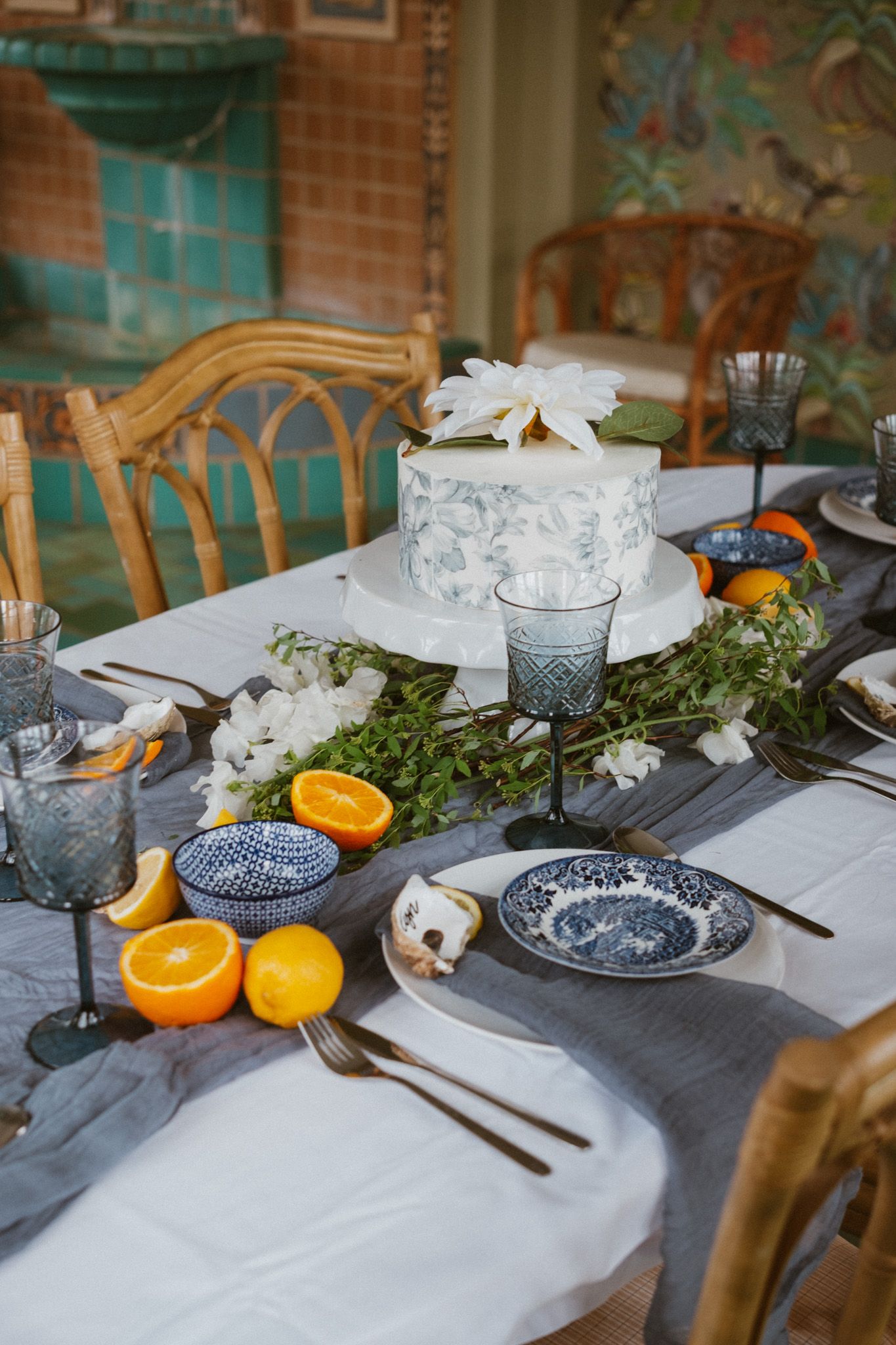 Wedding table with blue glassware, white cake, and orange slices on gray linen.