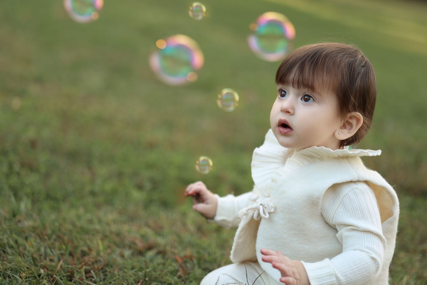 Portrait of a baby in soft white outfit sitting in a grassy field looking at iridescent floating bubbles