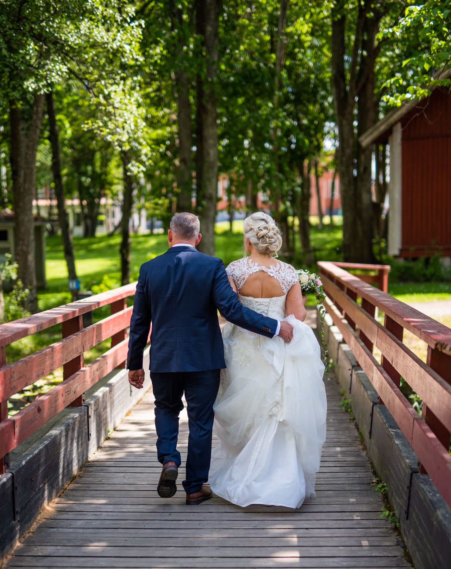 Bride and groom walking hand in hand across a wooden bridge surrounded by lush green trees.