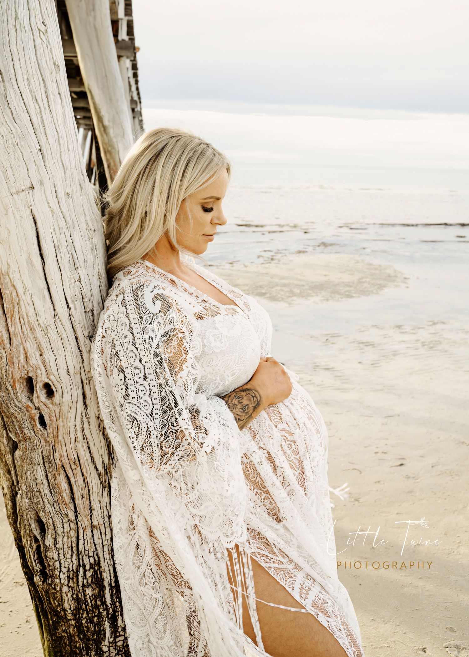 Pregnant woman in white lace kimono leaning against driftwood on a serene beach, cradling her baby bump.
