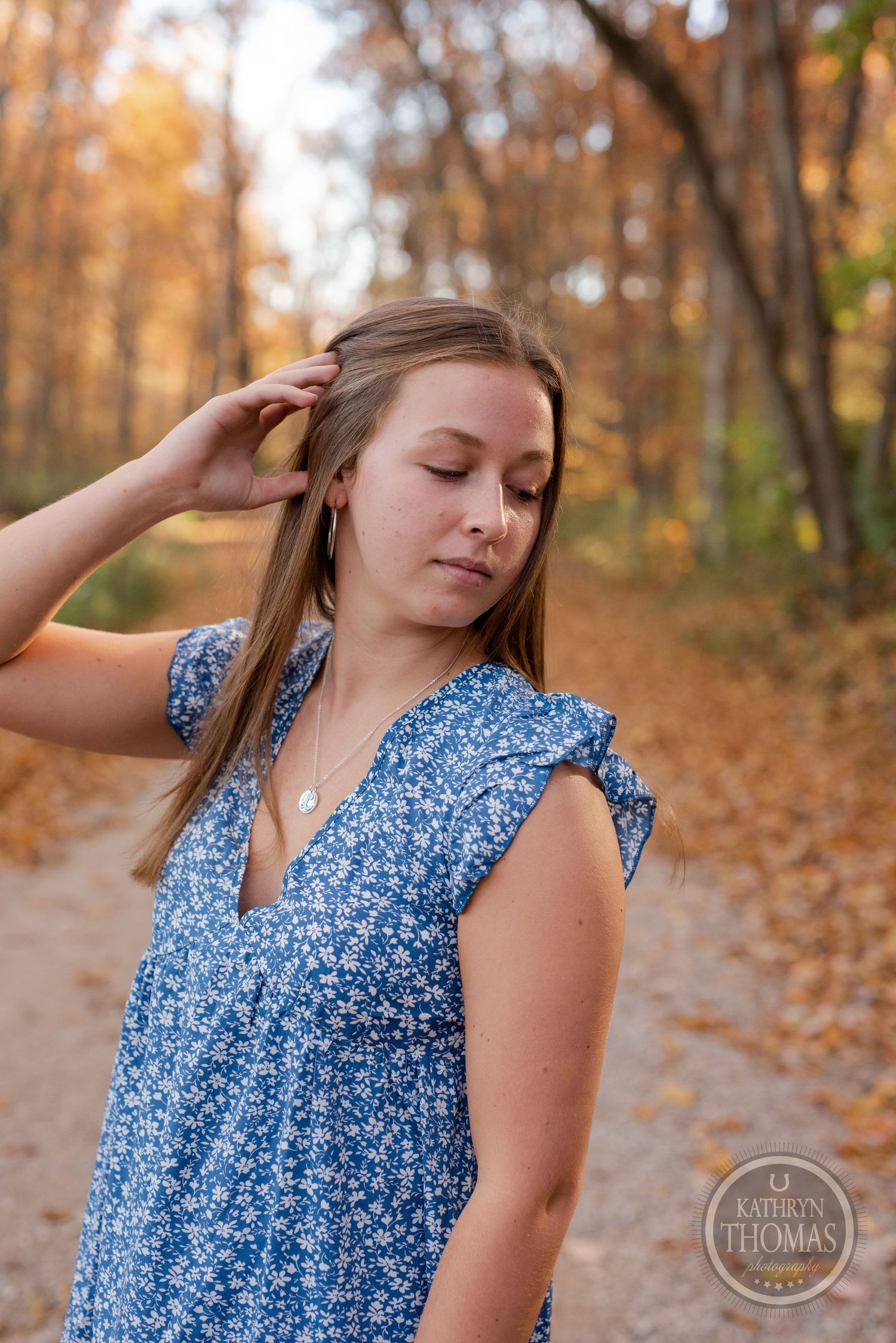 Someone in a blue patterned dress touches their hair while standing on an autumn forest path.