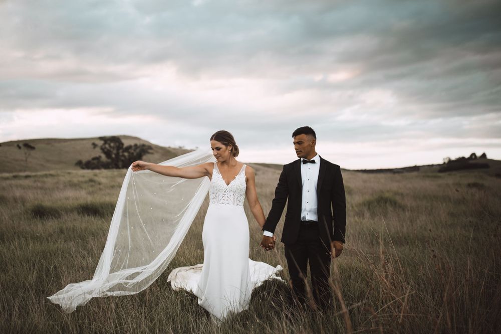 Wedding couple dances playfully in an open field with flowing veil and dramatic sky.
