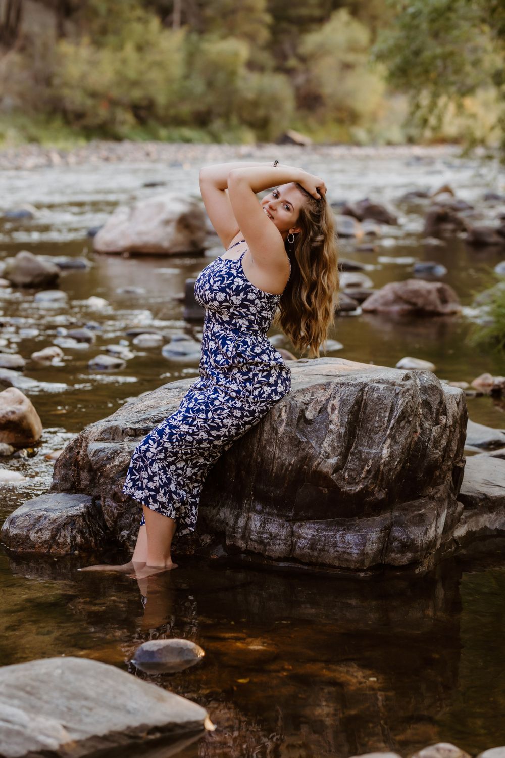 A senior girl in a floral dress poses on rocks in a serene river setting during golden hour.