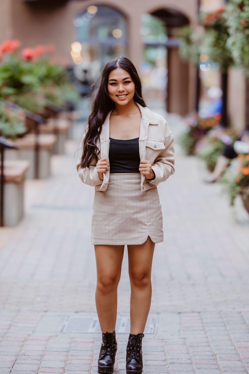 Senior girl in beige blazer and matching skirt poses on cobblestone street with flowers and urban backdrop in downtown Fort Collins.