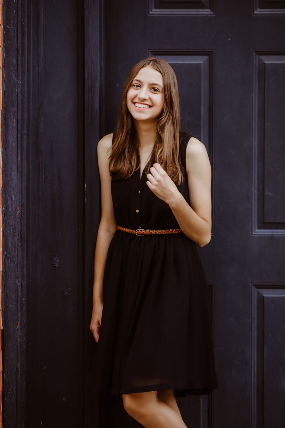A senior girl in a black sleeveless dress with a brown belt poses against a dark blue door, smiling warmly.