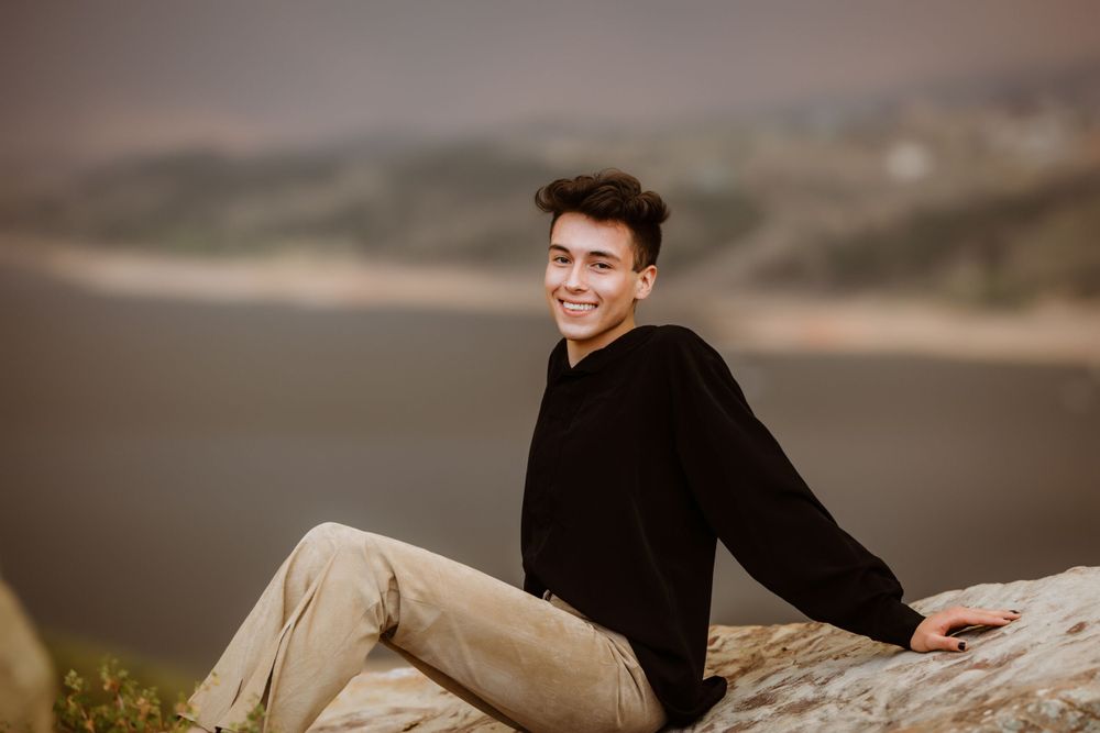 A senior boy in a black sweater sits on a rock formation during golden hour with a blurred natural landscape background at Horsetooth.