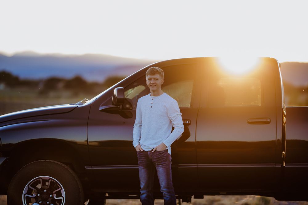 A senior boy stands in front of a black pickup truck at sunset with warm golden light illuminating the scene.