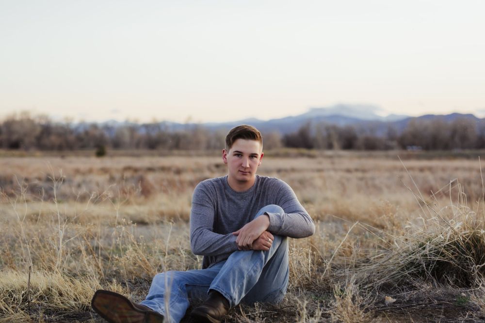 A senior boy sits in a field wearing a gray sweatshirt with mountains visible in the background at sunset.