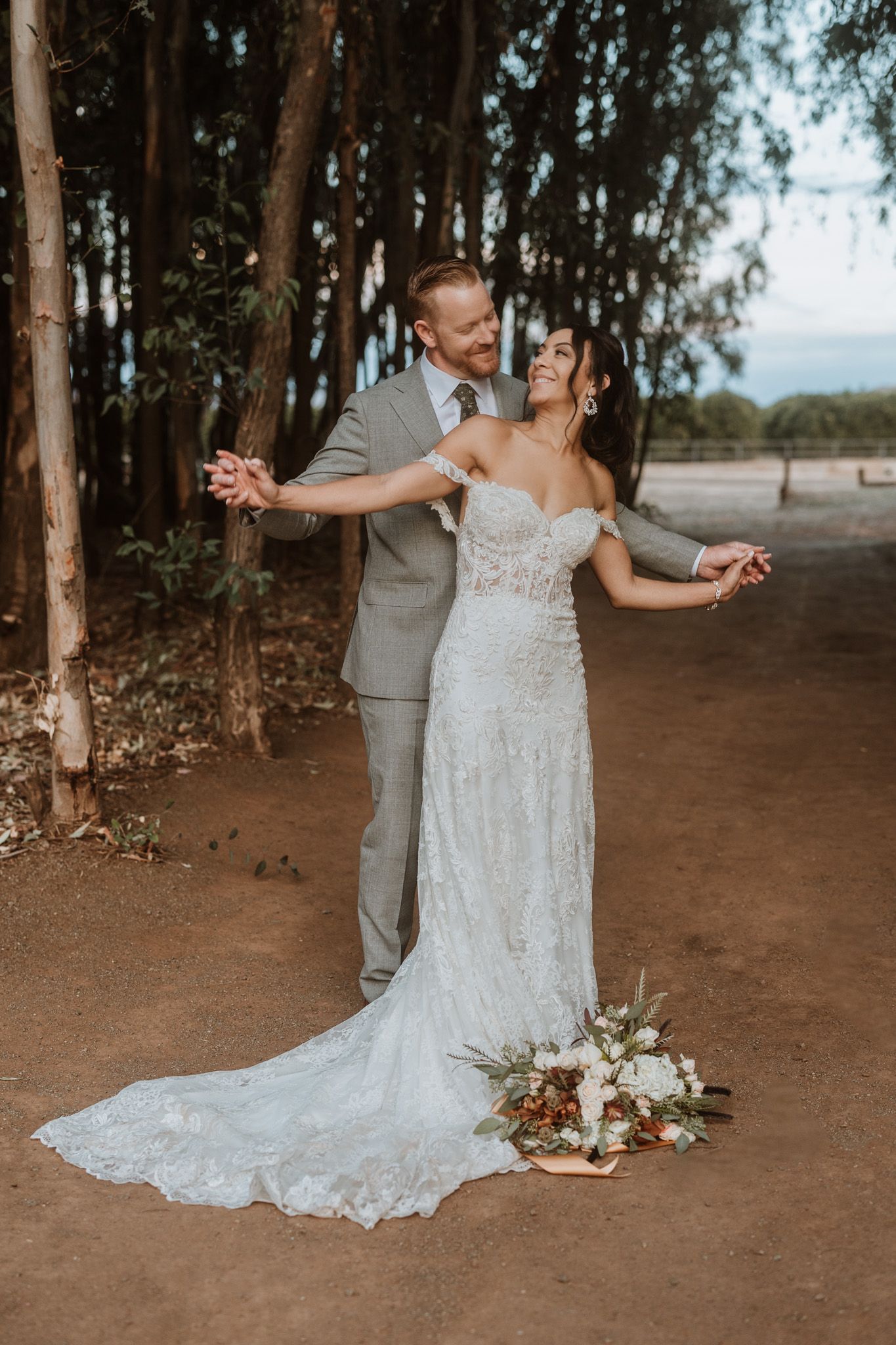 Romantic newlyweds dance on dirt path among eucalyptus trees with white lace wedding gown and gray suit.