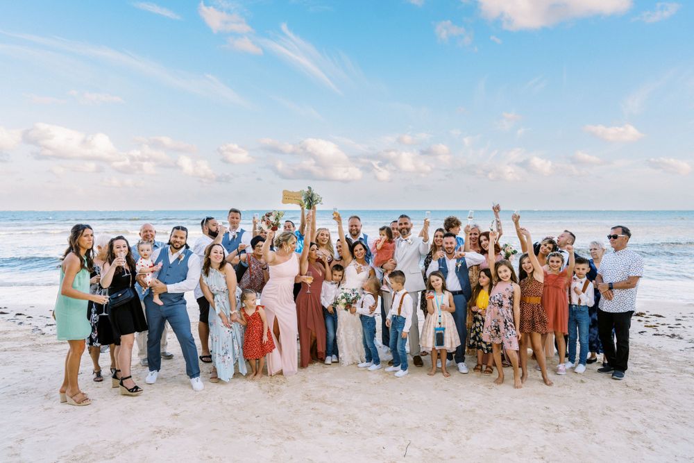 Large wedding party celebrating on a sunny beach with ocean waves in the background.