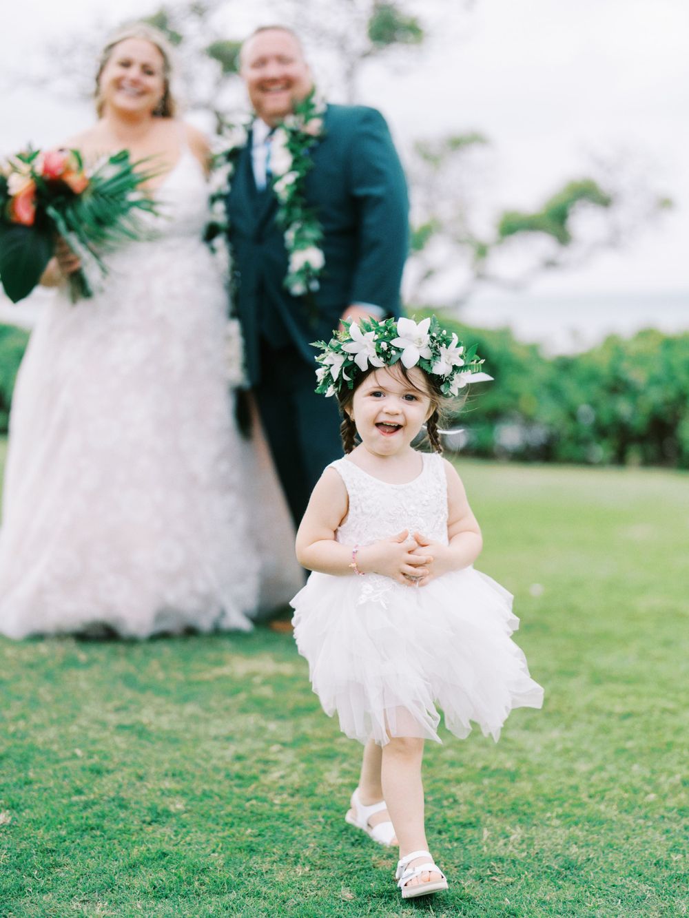 A young flower girl in a white dress runs joyfully across a green lawn during a wedding.