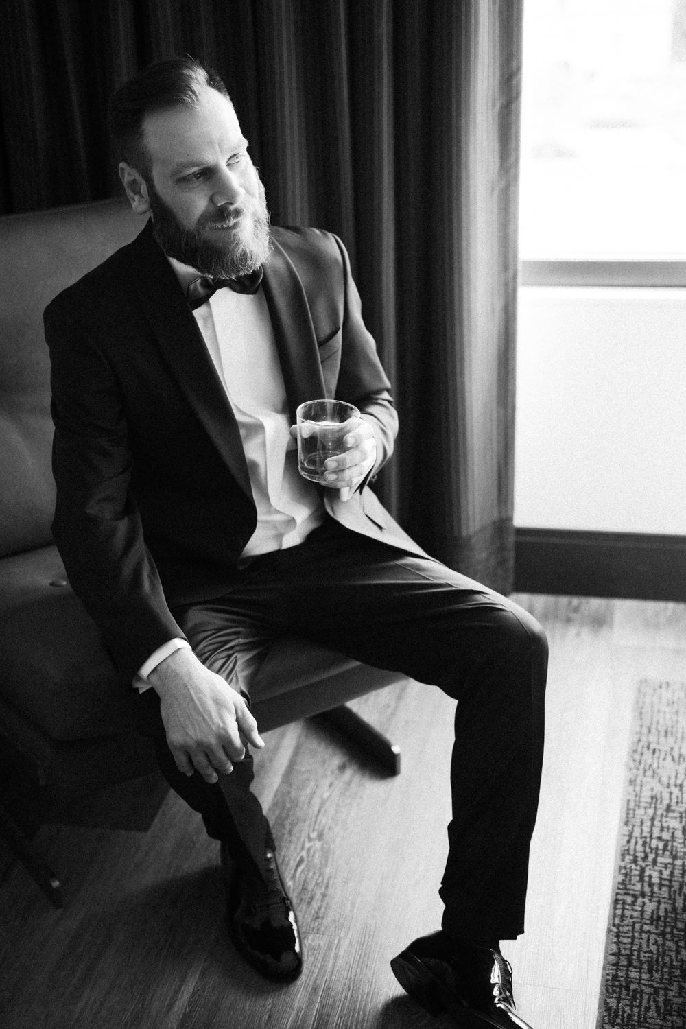 Black and white photo of person in tuxedo sitting by window with drink in hand.