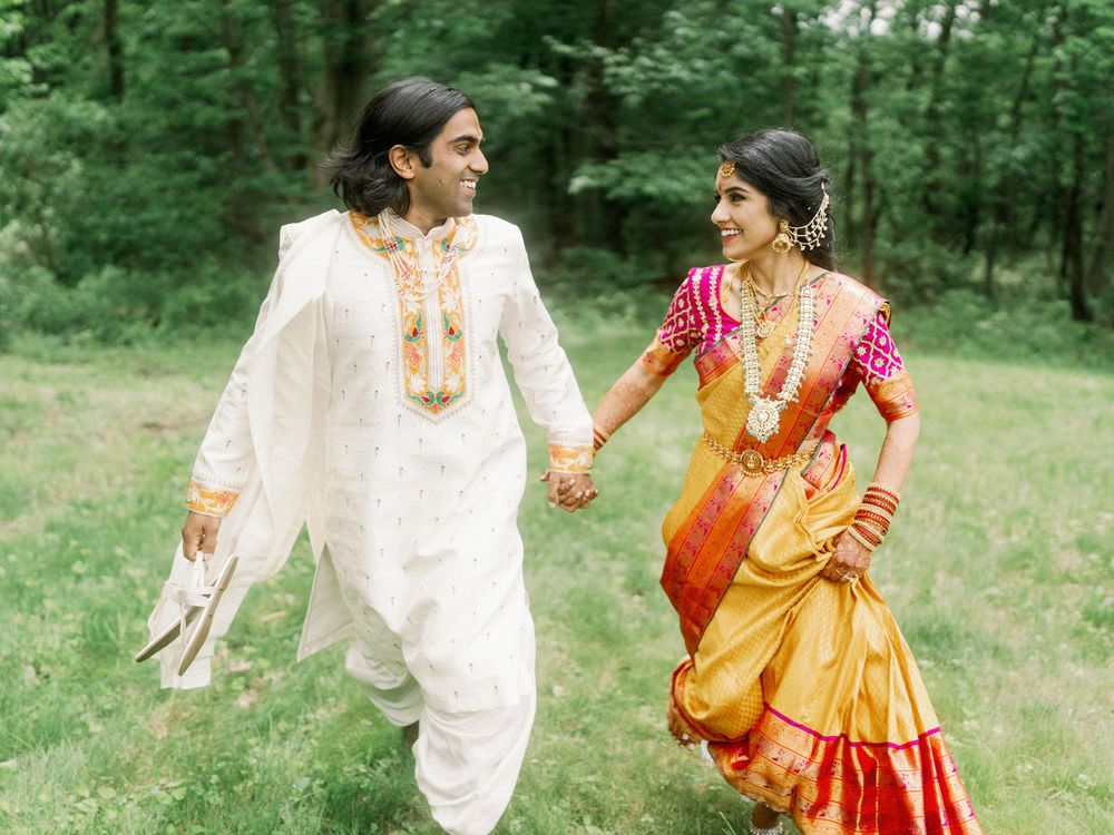 Couple in traditional Indian wedding attire running playfully through a green outdoor setting.